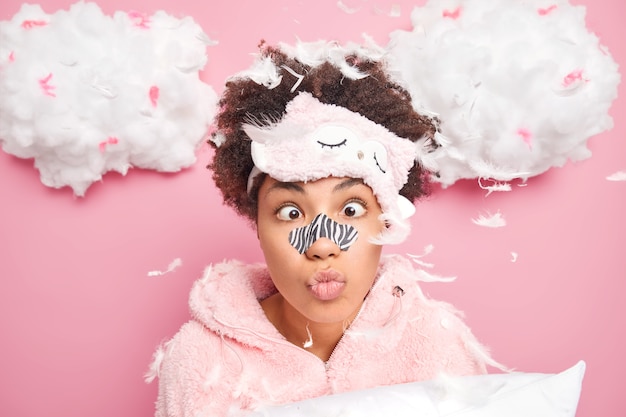 Mulher afro-americana engraçada faz careta tem aplicador de máscara no nariz cruza os olhos passa por procedimentos de beleza para pele macia e saudável vestida em pijamas posa com travesseiro com penas ao redor