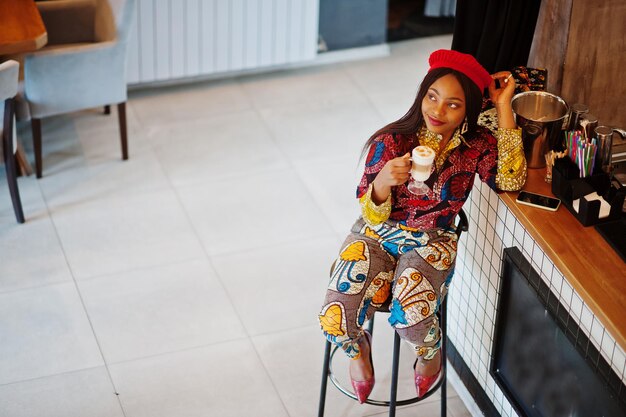 Mulher afro-americana entusiasmada com roupa colorida da moda com boina vermelha relaxando no café aconchegante sentado no balcão do bar