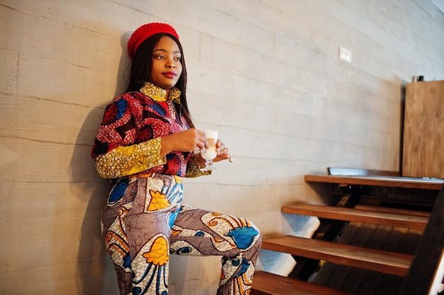 Mulher afro-americana entusiasmada com roupa colorida na moda com boina vermelha relaxando no café aconchegante em pé nas escadas com um copo de bebida quente nas mãos