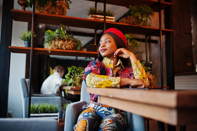 Mulher afro-americana entusiasmada em roupas coloridas da moda com boina vermelha relaxando no café aconchegante