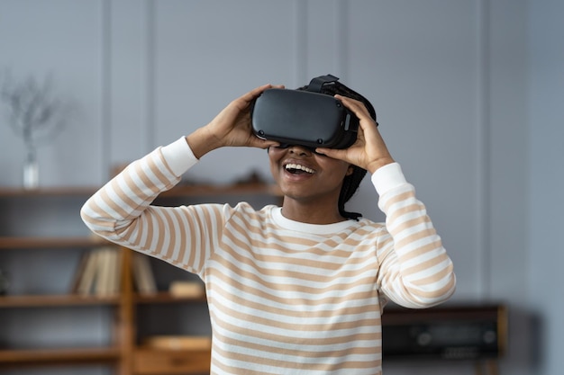 Mulher afro-americana espantada testando capacete VR moderno de alta tecnologia obtendo experiência 3D em casa