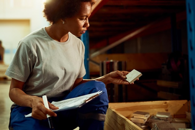 Mulher afro-americana examinando produtos acabados e escrevendo notas enquanto trabalhava em um armazém