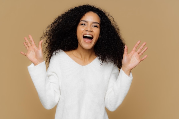 Mulher afro-americana feliz emocional com cabelo preto nítido levanta gestos de mãos observa ativamente algo maravilhoso e incrível usa modelos de jumper branco macio sobre fundo marrom