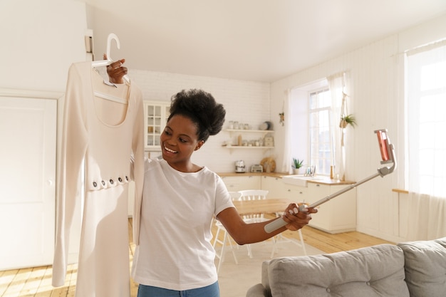 Mulher afro-americana feliz mostrando vestido de verão da moda ao lado do celular no tripé online em casa