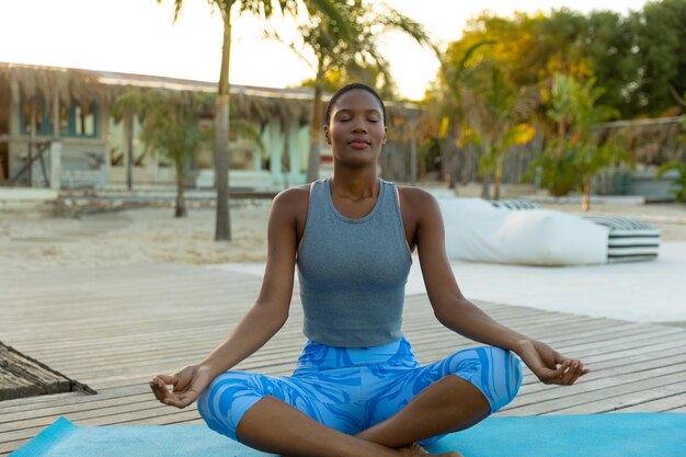 Mulher afro-americana feliz praticando meditação yoga sentada na praia ensolarada. Posição de lótus, estilo de vida saudável, bem-estar, relaxamento, verão e férias, inalterados.