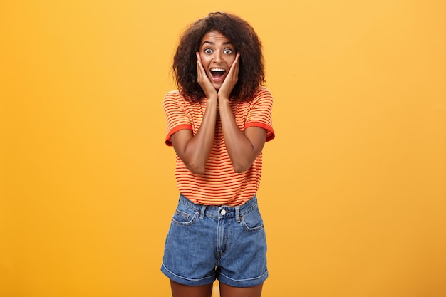 Mulher afro-americana impressionada, apoiando o rosto nas palmas das mãos, sorrindo amplamente com uma expressão divertida.