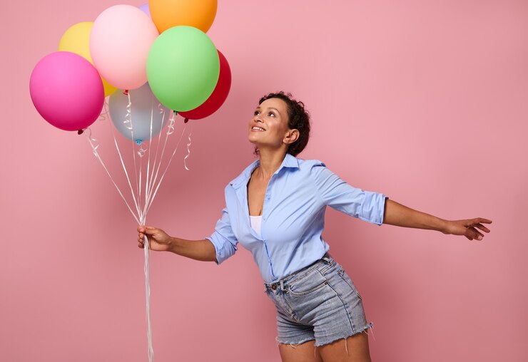 Foto mulher afro-americana jovem e feliz, sorridente, de raça mista, com um monte de balões coloridos do arco-íris olhando para cima e se divertindo, vestindo shorts jeans e camisa azul no fundo da parede rosa com espaço de cópia