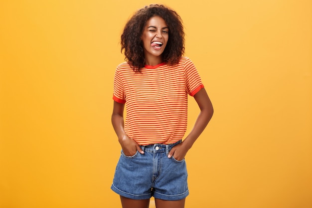 Mulher afro-americana jovem entusiasmada alegre com penteado afro, piscando alegremente, mostrando a língua.