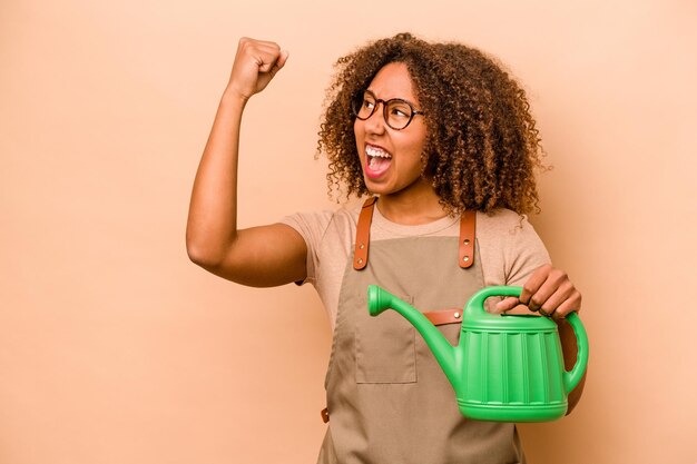 Mulher afro-americana jovem jardineiro segurando irrigação isolada em fundo bege, levantando o punho após um conceito de vencedor da vitória