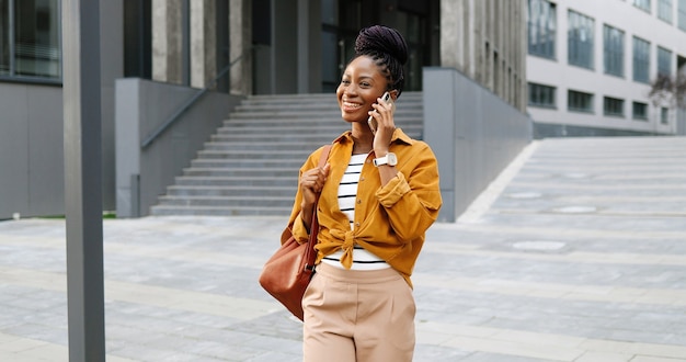 Mulher afro-americana linda feliz sorriu falando no celular e andando lá fora. Mulher alegre falando no celular e passeando. Conversa telefônica.