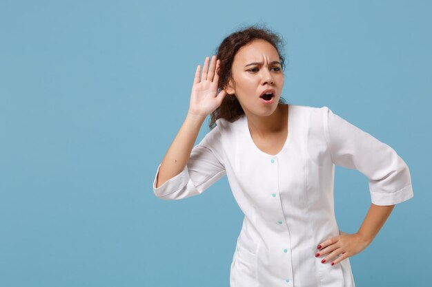 Mulher afro-americana médica isolada em fundo azul. Médica em vestido médico branco tentando ouvi-lo com a mão perto da orelha. Conceito de medicina de saúde de pessoal de saúde. Simule o espaço da cópia.