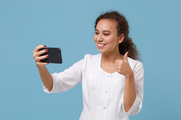 Mulher afro-americana médica isolada em fundo azul. médico em vestido médico fazendo selfie filmado no celular faz videochamada aparecendo o polegar. conceito de pessoal de saúde. simule o espaço da cópia.