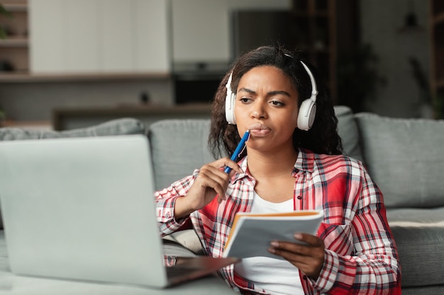 Foto mulher afro-americana milenar séria em fones de ouvido sem fio estudando com bloco de notas e laptop