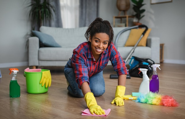 Mulher afro-americana milenar sorridente em luvas de borracha lavando o chão com material de limpeza