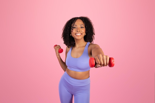 Mulher afro-americana milenar sorridente em roupas esportivas fazendo exercícios manuais com halteres