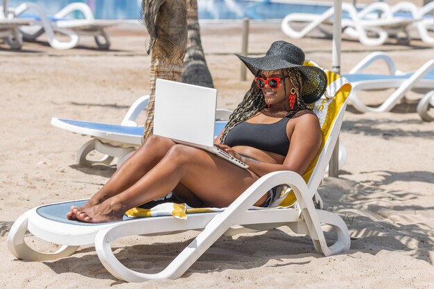 Mulher afro-americana na praia relaxando na espreguiçadeira usando o laptop. Mulher adulta alegre, aproveitando um dia de verão na praia.