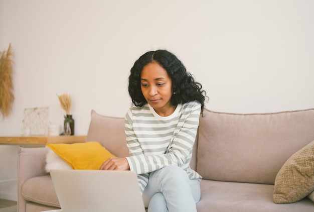 Mulher afro-americana navegando laptop enquanto está sentado no sofá usando dispositivo digital para trabalho e estudo