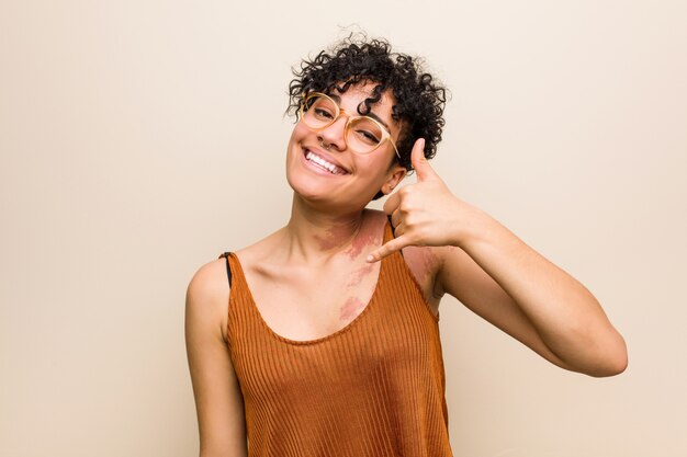 Mulher afro-americana nova com a marca de nascimento da pele que mostra um gesto de chamada de telefone móvel com os dedos.