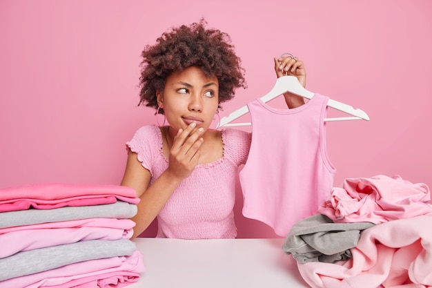 Mulher afro-americana pensativa segurando a camisa no cabide desvia o olhar com expressão pensativa e se senta à mesa com pilhas de roupas desdobradas isoladas sobre rosa