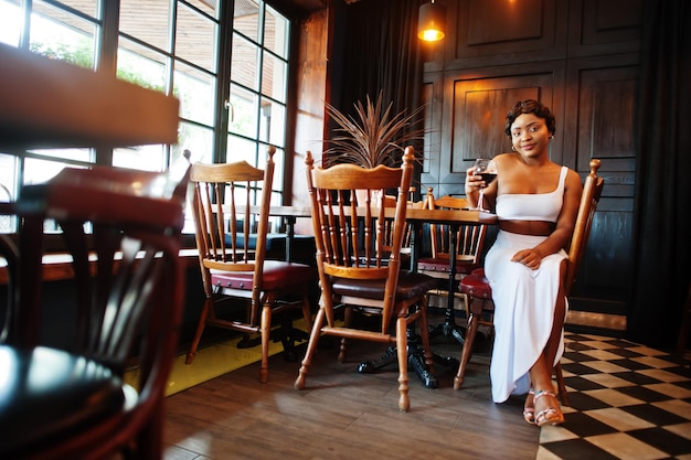 Mulher afro-americana, penteado retrô em vestido branco no restaurante com um copo de vinho.