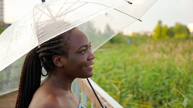 Mulher afro-americana positiva com grande guarda-chuva transparente em pé na ponte de madeira vazia contra juncos verdes vista de perto