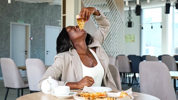 Mulher afro-americana positiva com jaqueta cinza gosta de comer pizza deliciosa à mesa com bule e xícara no café contemporâneo leve