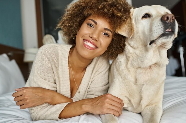 Mulher afro-americana positiva com seu labrador deitado em uma cama em um hotel de viagens para animais de estimação