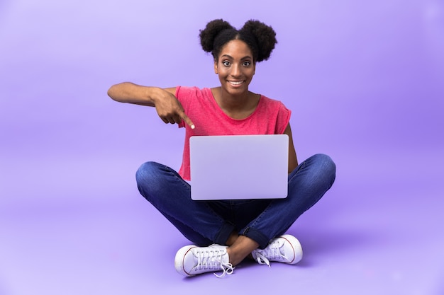 mulher afro-americana positiva sorrindo e usando laptop prateado, sentado no chão com as pernas cruzadas, isolado