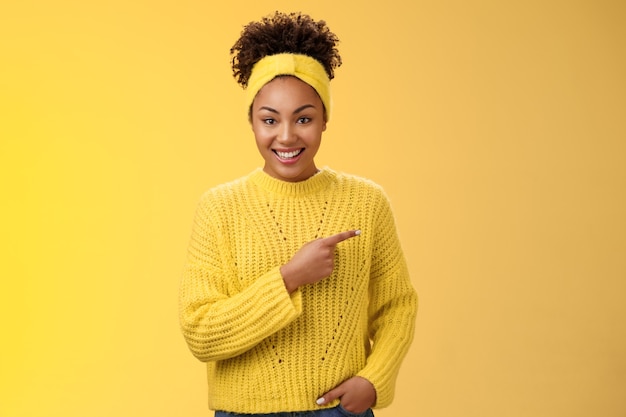 Mulher afro-americana relaxada amigável ougoing casualmente apontando para a direita durante a conversa sobre o recente novo café aberto promoções de descontos incríveis em pé feliz fundo amarelo. Copie o espaço