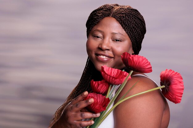 Mulher afro-americana segurando o retrato de flores de papoula, mostrando o conceito de feminilidade e ternura. Atraente modelo plus size posando com lindas plantas de verão em estúdio de perto