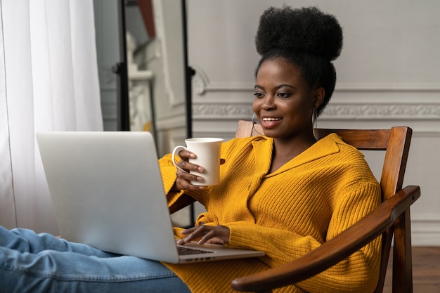 Mulher afro-americana sentada na cadeira, assistindo ao webinar, trabalhando online no laptop, conversando no chat por vídeo, bebendo chá ou café.
