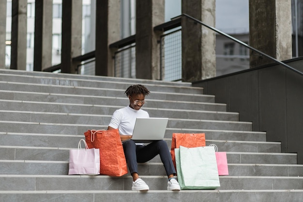 Mulher afro-americana sentada nos degraus do shopping com um laptop e sacolas coloridas