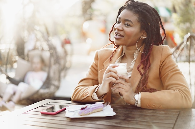 Foto mulher afro-americana sorridente, bebendo café e comendo sobremesa em uma cafeteria ao ar livre