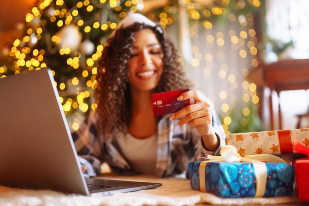 Mulher afro-americana sorridente usando laptop sentado perto da árvore de Natal com cartão de compras on-line