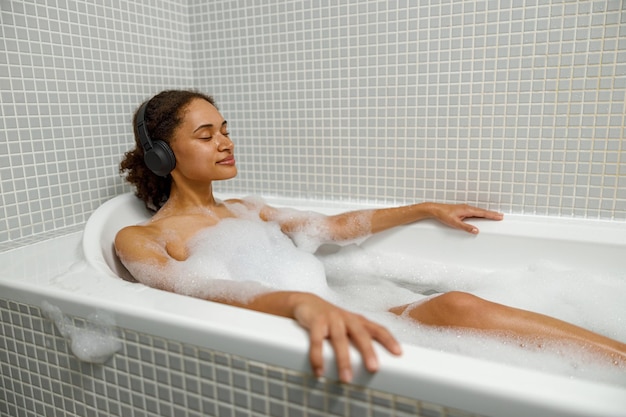 Foto mulher afro-americana toma banho de espuma na banheira e ouve música conceito de beleza e higiene