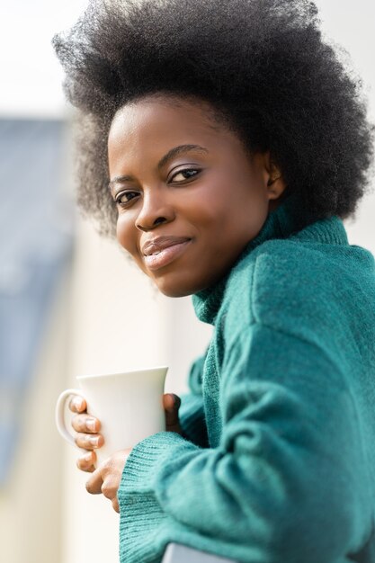 Mulher afro-americana tomando uma xícara de chá ou café olhando para a câmera em pé na varanda