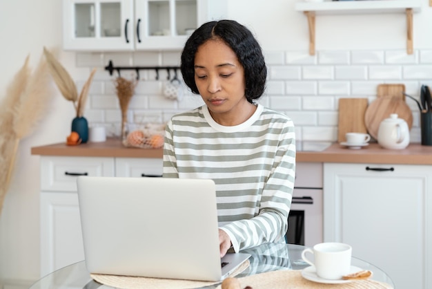 Mulher afro-americana usando laptop enquanto está sentado na cozinha confiante e focado no trabalho remoto