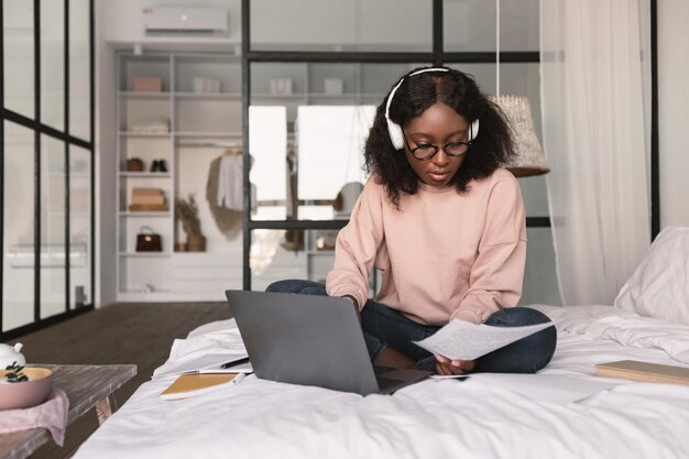 Mulher afro-americana usando laptop lendo papel sentado no quarto