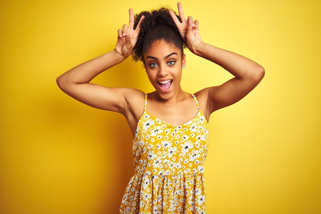 Mulher afro-americana usando vestido floral casual em pé sobre fundo amarelo isolado Posando engraçado e louco com os dedos na cabeça como orelhas de coelho sorrindo alegre