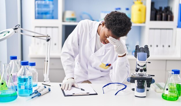 Mulher afro-americana vestindo uniforme de cientista estressada trabalhando no laboratório