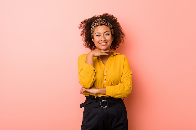 Mulher afro-americano nova contra um sorriso cor-de-rosa da parede feliz e confiante, tocando o queixo com mão.