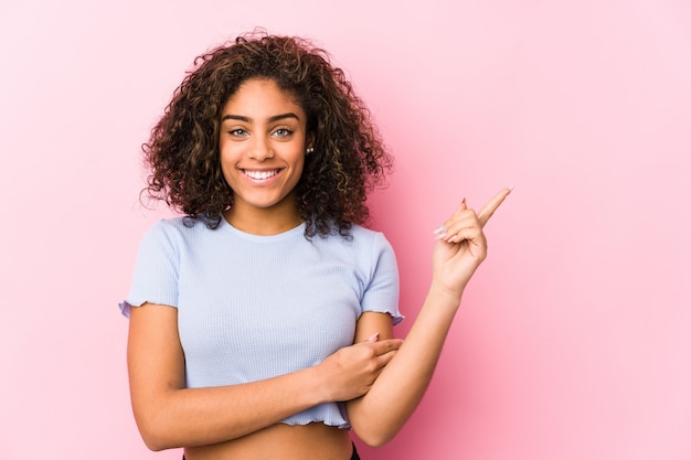 Foto mulher afro-americano nova contra uma parede cor-de-rosa que sorri alegremente apontando com o dedo indicador afastado.