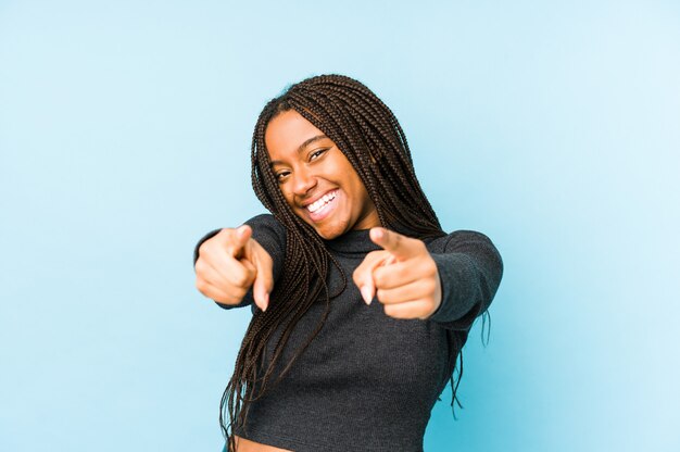 Mulher afro-americano nova isolada em sorrisos alegres da parede azul que aponta à parte dianteira.