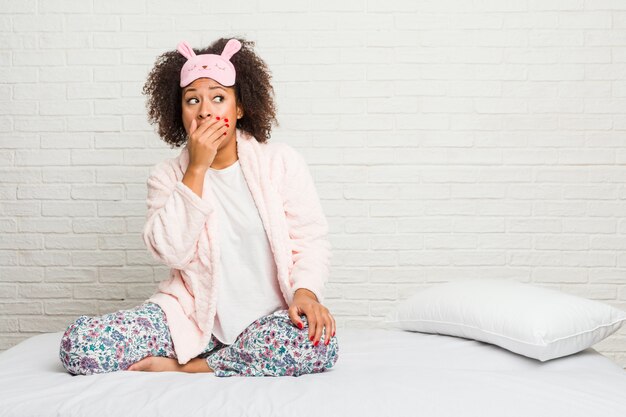 Mulher afro-americano nova na cama que veste o pijama pensativo olhando a um espaço da cópia que cobre a boca com a mão.