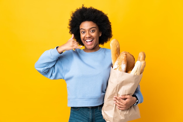 Mulher afro-americano nova que compra algo pão na parede amarela que faz o gesto do telefone. ligue para mim de volta