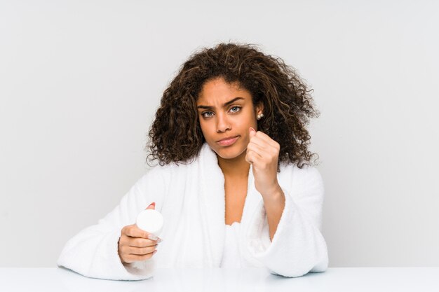 Mulher afro-americano nova que guarda um creme hidratante que mostra o punho à câmera, expressão facial agressiva.