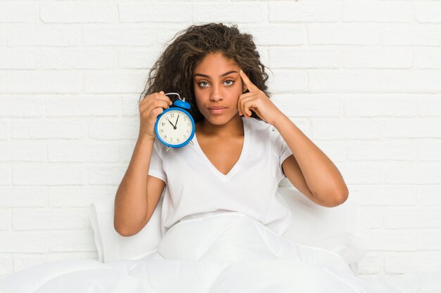 Mulher afro-americano nova que senta-se na cama que guarda um despertador que aponta seu templo com o dedo, pensando, focalizado em uma tarefa.