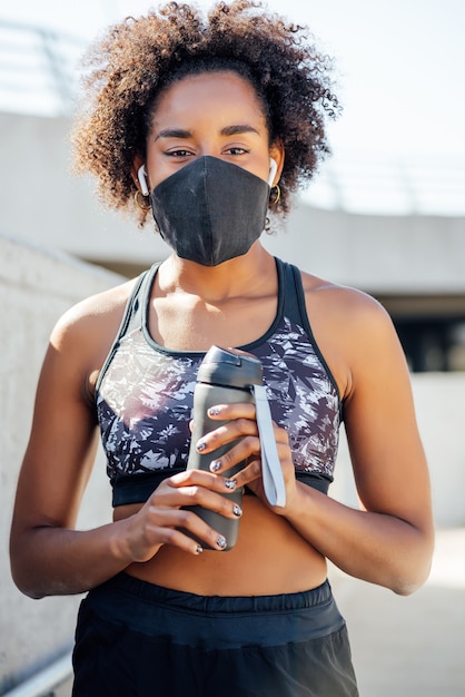 Mulher afro-atlética usando máscara facial e segurando uma garrafa de água depois do treino ao ar livre. esporte e estilo de vida saudável.