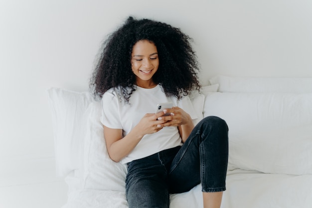 Mulher afro bonita relaxada com expressão alegre usa telefone celular moderno, navega na internet, posa em cama confortável branca, conectada ao wifi, navega na página da web, desfruta de atmosfera doméstica