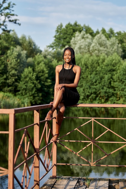 Foto mulher afro elegante em vestido de moda fica perto de um lago azul em um dia ensolarado de verão